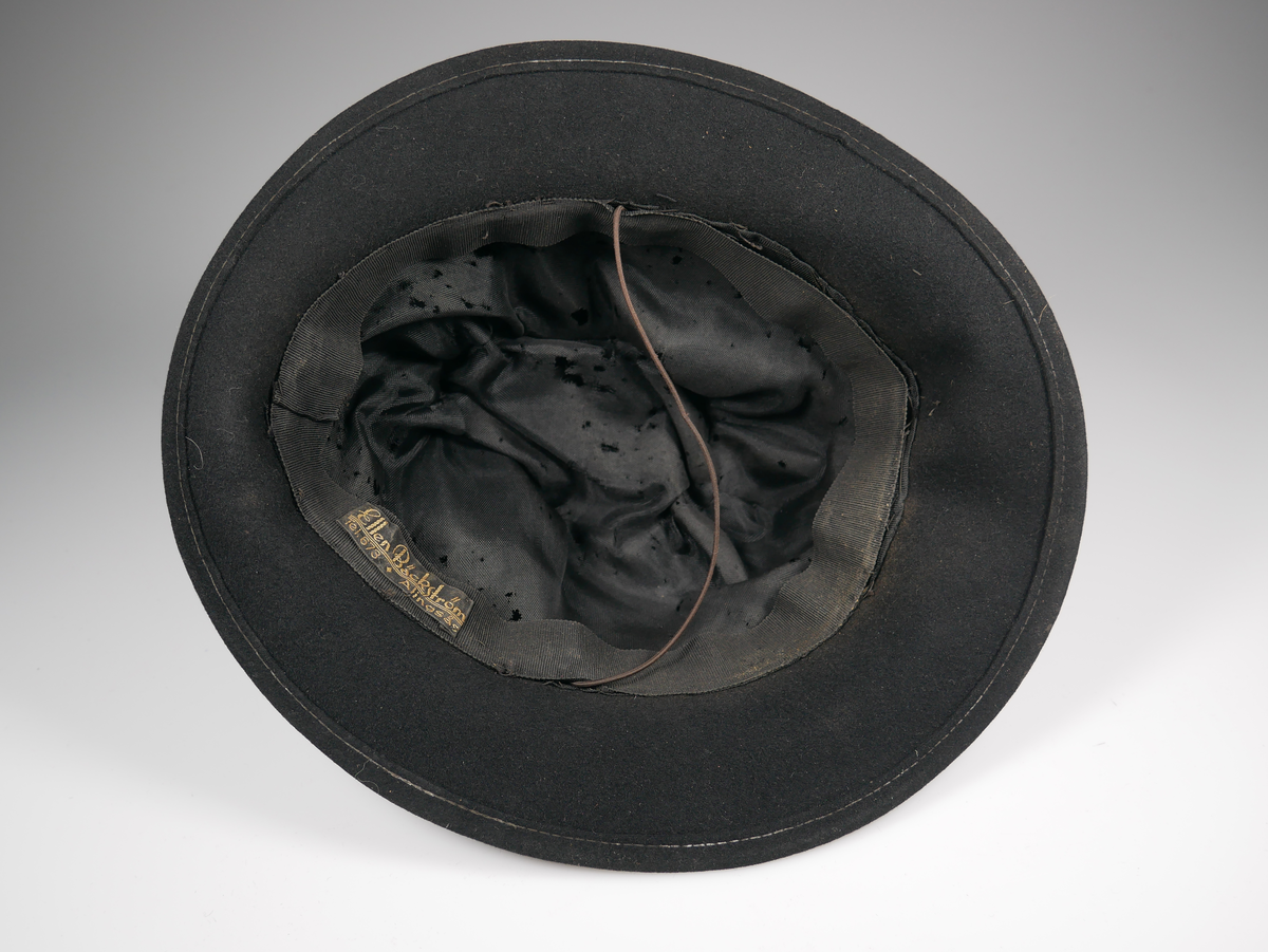 Svart damhatt av fedoramodell. Av filt. I hatten finns en etikett från "Ellen Bäckström Tel.673 Alingsås". 
Fodret mycket trasigt.