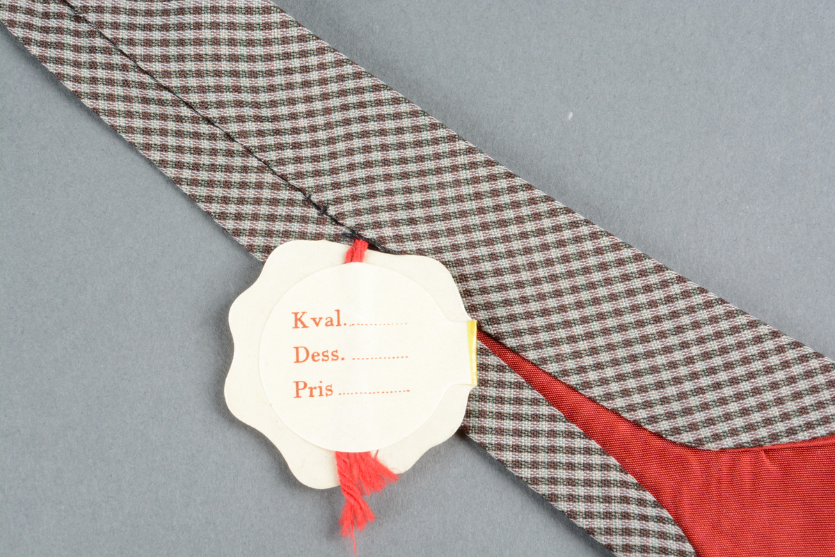 Slips av perlon og cuprama med brunt rutete skall (konvolutt).  Rødt fôr. Ingen rem for å holde slipsets bakstykke på plass ved bruk. 45 graders søm på bakstykket hvor delene er sydd sammen.