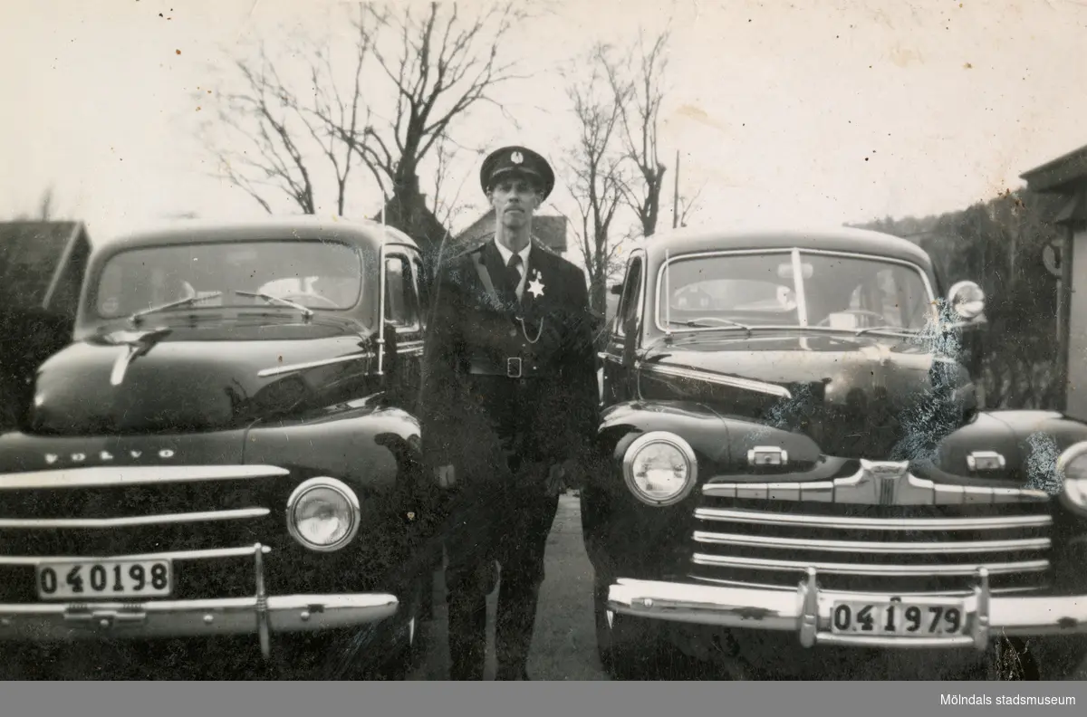Fritiof (elev boende på Stretereds skolhem) iförd polisuniform och skärmmössa, står mellan två taxibilar vid Kållered station 1940-tal. Han fick "hjälpa" polisen ibland. Taxistationen skymtas till vänster.