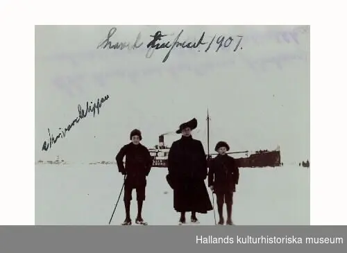 "Havet tillfruset 1907" har någon skrivit på denna bild av en kvinna och två pojkar som står på isen, med ett fartyg i bakgrunden och Skrivareklippan till vänster.