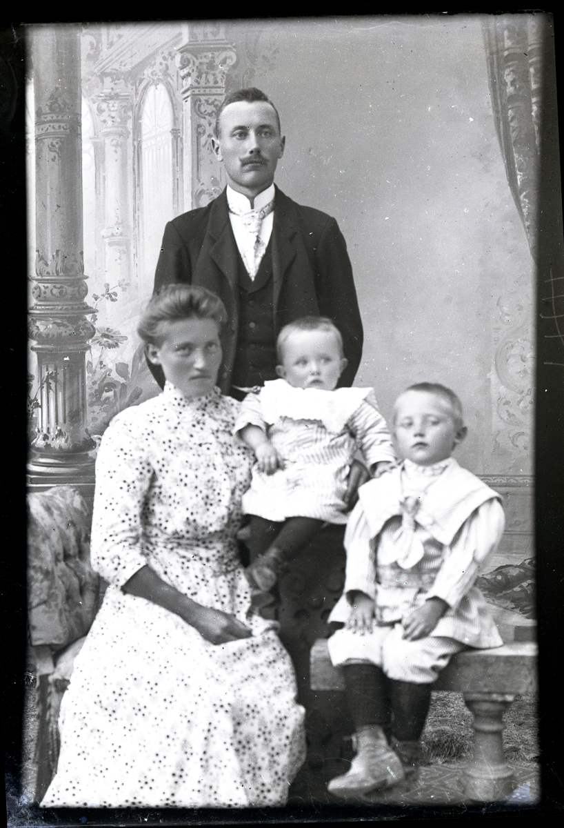 Atelierfoto. Familiebilde. En kvinne med to små barn sitter og en mann står bak.