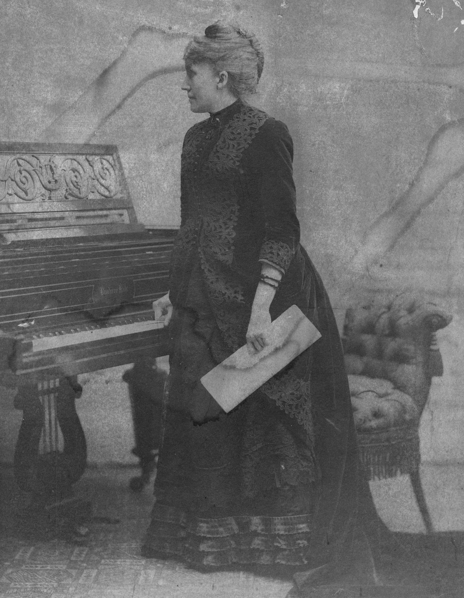 Agathe Backer i kjole med kapper og blonder ved et lite flygel. 1900