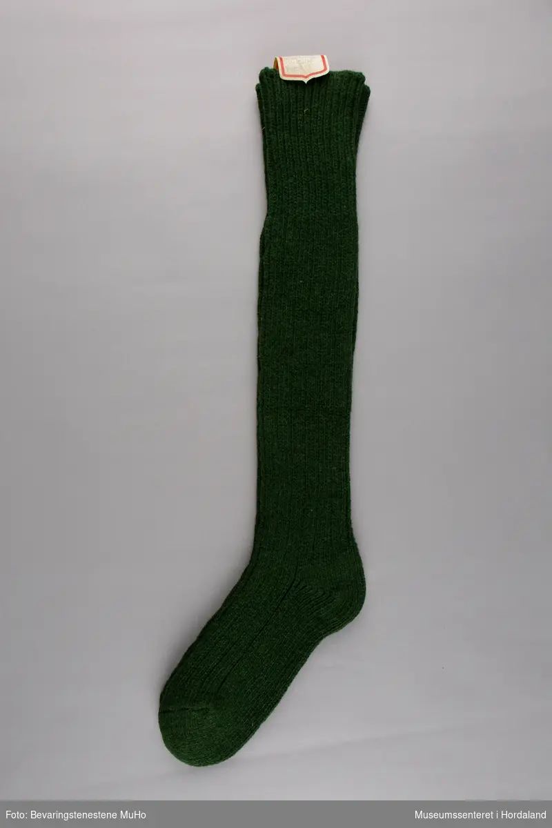 Eit par mørk grøne Krone ribbestrikka knelange ullstrømper. Strømpene er festa saman i eit par med ein klistrelapp øvst.