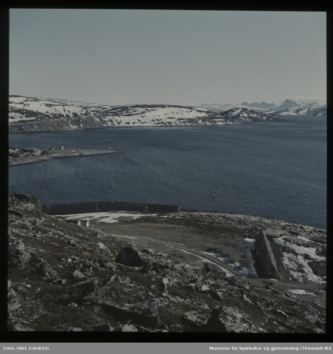 Bildet er fotografert fra Vardfjellet ovenfor Meland. I forgrunnen ser man fiskehjeller der fisken henger til tørk. Utsikten går over Rossmollbukta og innseilingen til Hammerfest havn til veien på Salenfjellet opp til Storsvingen og Rypefjellet i bakgrunn. Det ligger fortsatt snø.