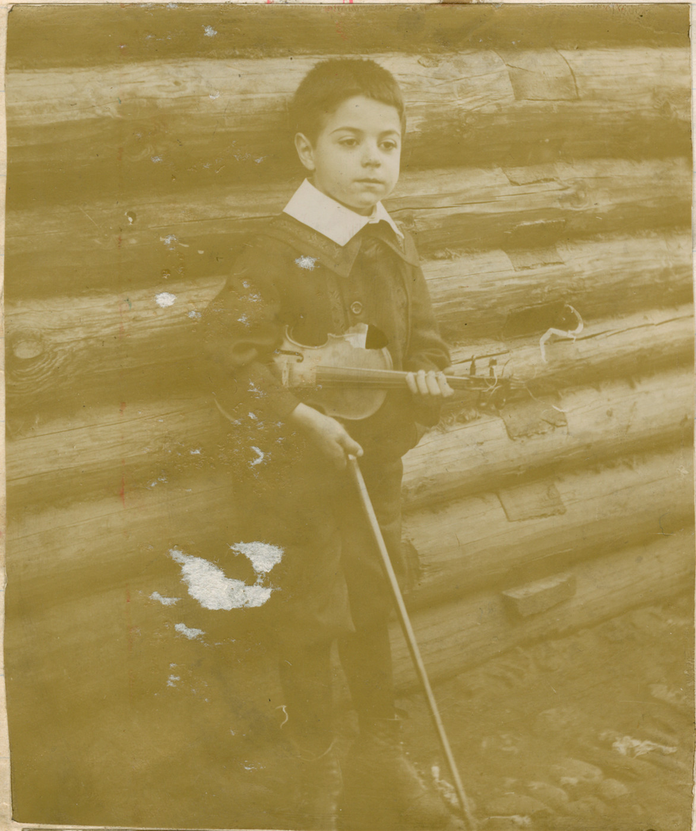 En gutt står lent mot en tømmerbygning. Han holder en fiolin.