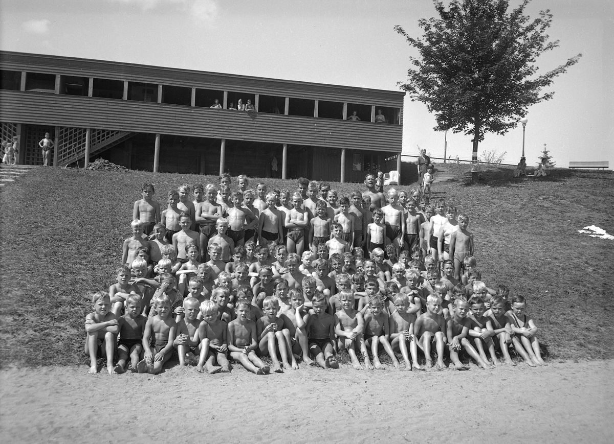 En pojkgrupp med simskoleelever har samlats i slänten nedanför Tinnerbäcksbadets barnavdelning. Högtidligt tittar de mot fotograf Sven Källbergs kamera. Uppe till höger ses den legendariske simläraren Victor Lundkvist. Året är 1945.