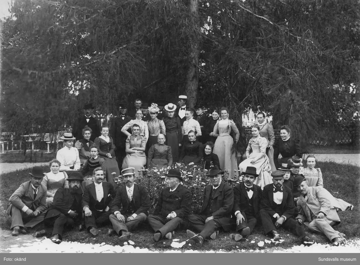 Gruppfoto med människor som sitter och står i en gräsrundel i en park. Eventuellt är bilden från sommarrestaurangen Tivoli. På baksidan står: S-vall. Ur Anders Nylanders samling.