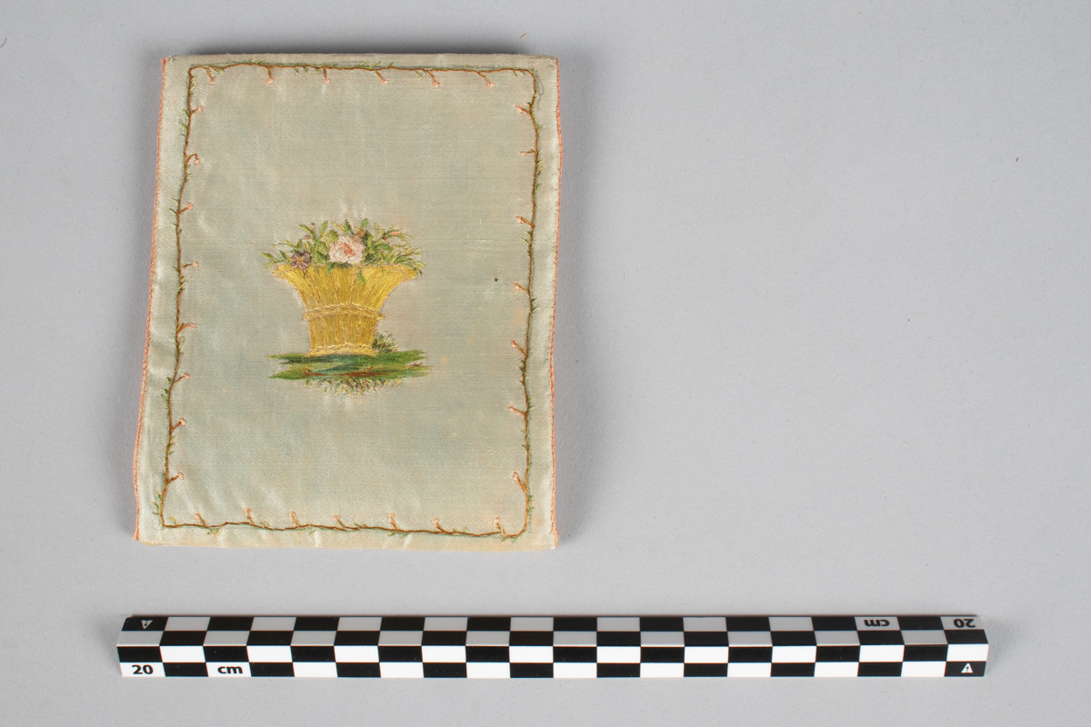 Brodert med border. Blomster og vase i grønt, hvitt, rødt, brunt og gult. Monogram N brodert inni. Utenpå er brodert 1800.