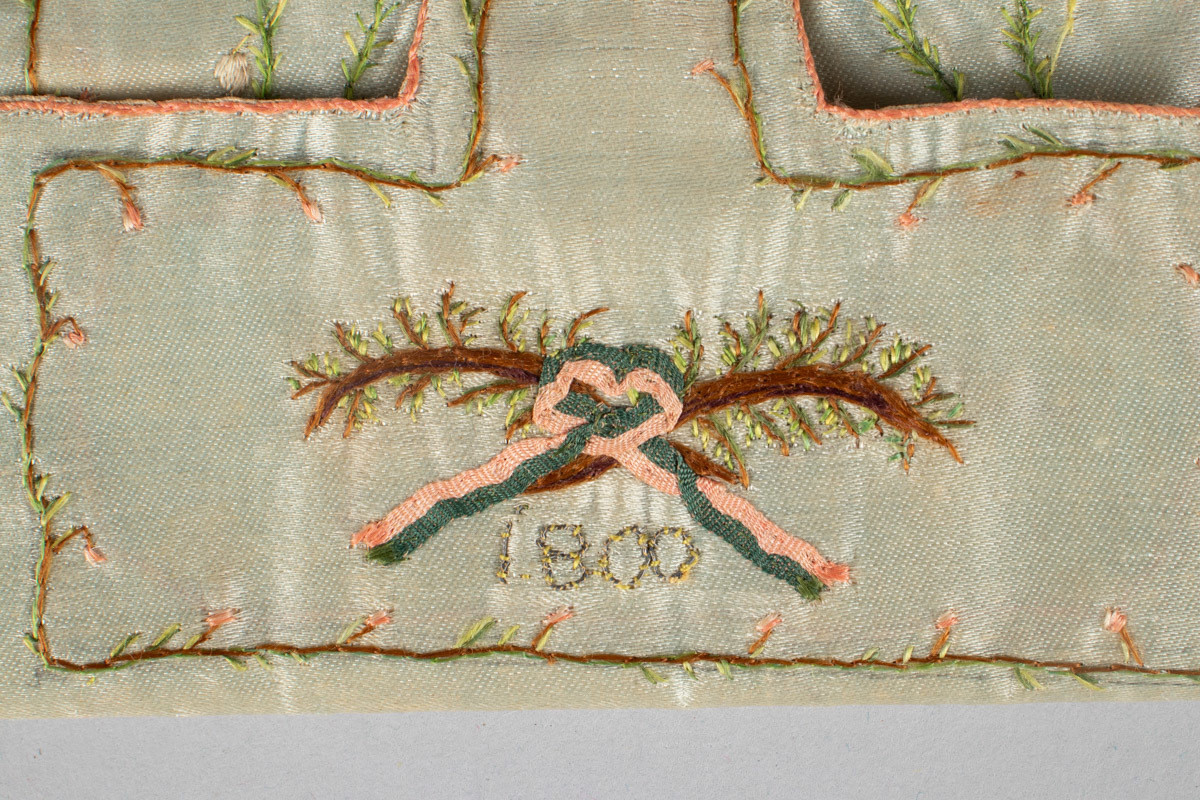 Brodert med border. Blomster og vase i grønt, hvitt, rødt, brunt og gult. Monogram N brodert inni. Utenpå er brodert 1800.