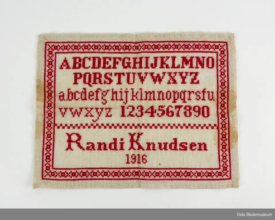 Rektangulær navneduk med røde sting. På navneduken er det brodert bokstaver og tall samt "Randi Knudsen" og "1916". Det er også brodert en pyntebord.