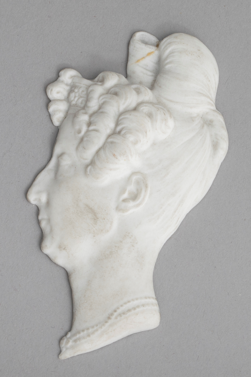 Kvinneportrett i uglasert porselen (biscuit). Kvinnehode i relieff, profil mot høyre, oppsatt hår med topp på issen, tinningskrøller og perlekjede rundt halsen. Portrettet er av dronning Marie-Amélie (1830-1848), Frankrike.