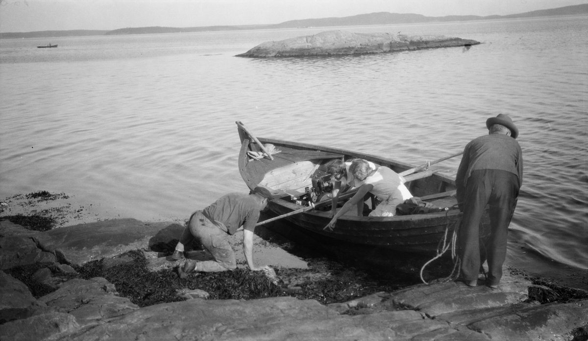 To kvinner og en mann oppdager noe i fjæra. En fjerde mann ordner med tauene til en trebåt. Antagelig et sted i skjærgården i Oslofjorden.