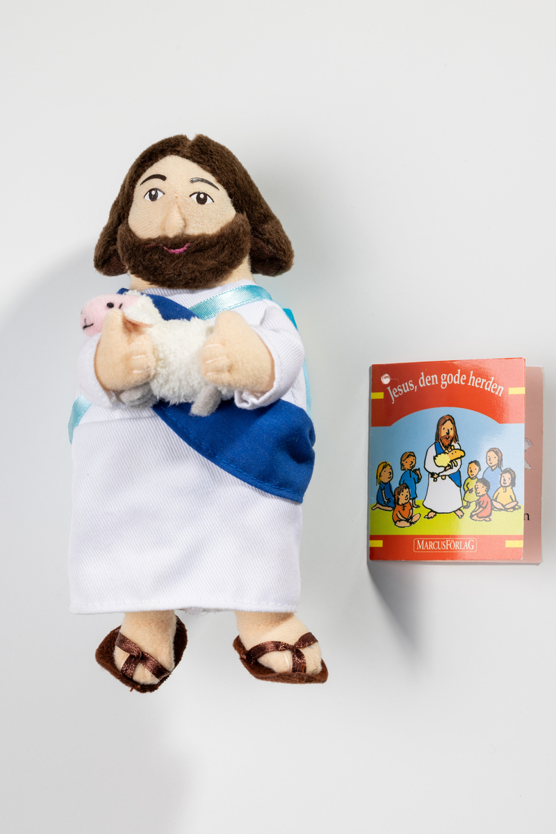 Docka, Jesusdocka med bok på ryggen, av textil. Jesus håller i ett lamm med båda händerna framför sig. Dockan har brunt hår och skägg, är iklädd en fotsid vit klädnad och har ett par bruna sandaler på fötterna. En turkos ryggsäck är fastsydd på kroppen med ett turkost band och ett brett blått band. När man öppnar ryggsäcken, med kardborreband, är där en liten bok om "Jesus den gode herden" illustrerad med teckningar.

Text av Stephanie St. Pierre, Illustrationer av Emily Bolam. Svensk text av Annica Hällzon. Bok nr 9453. ISBN 91 7999 4539. Copyright Bookworks LLC, England. Copyright Sverige Marcus Förlag, Box 1712, 70117 Örebro. Tel 019/165400, fax 019/120650, e-mail marcus@express.se, Made in China.