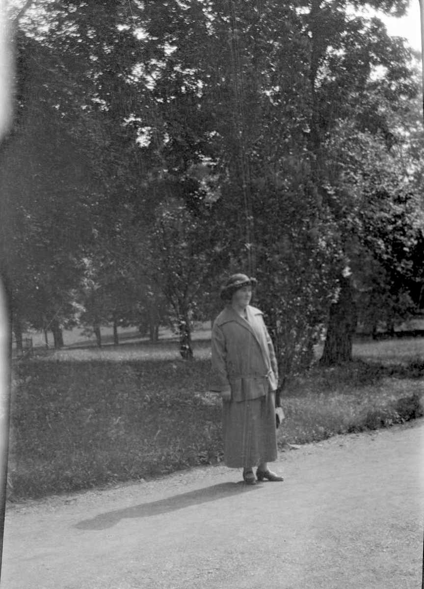 Landskap
Dame med drakt og hatt i en park med store trær.
