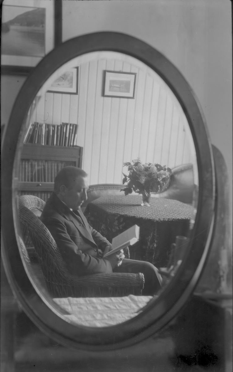 Portrett av mann som sitter i godstolen og leser i ei bok. Bildet er tatt via et speil inne i stua.
