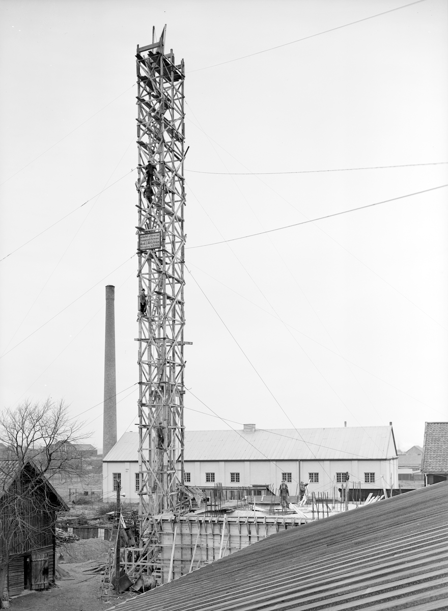 Byggarbetsplats vid Nykvarn i Linköping 1938. Närmare bestämt uppförandet av Lantmännens anläggning utmed nuvarande Nykvarnsgatan. Det till synes vanskligt konstruerade trätornet uppges vara hisstorn för att möjliggöra byggets fortskridan. Den på tornet monterade skylten upplyser oss att ansvarig byggnadsfirma var And. J. Ahlström från Helsingborg.