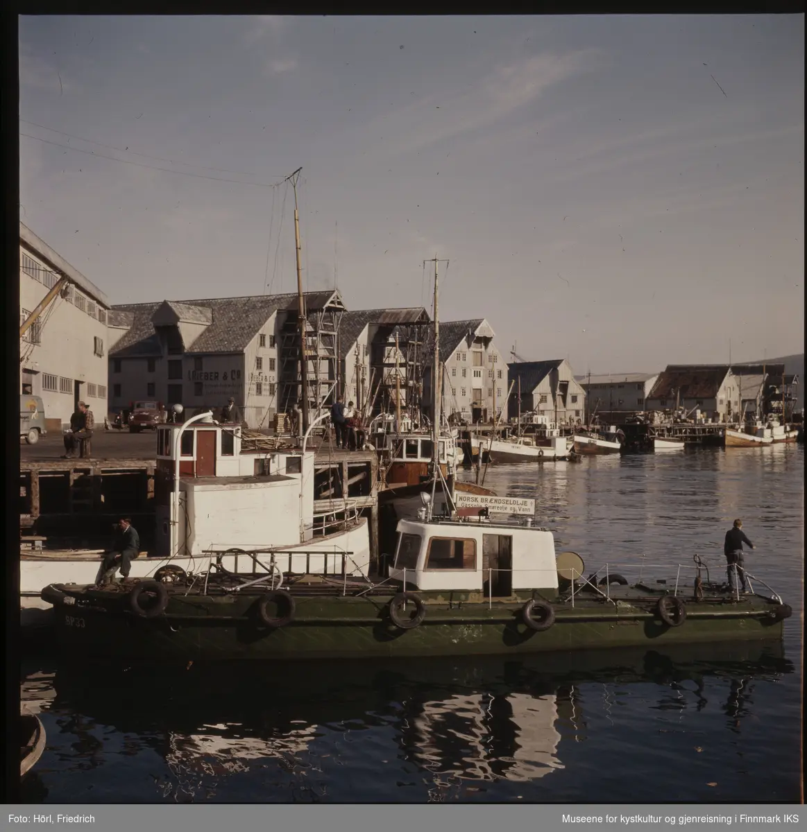 Havnesceneri i Tromsø i 1957 eller 1958. I forgrunnen ser man en grønn båt med et skilt for "Norsk Brændselsolje A/S". Ellers ser man fiskebåter ved kaia og mange pakkhus, blant annet til "Rieber & Co. A/S". Det ser ut som om det er flott vær og menn sitter på fiskebåten fremst i bilde samt på kaia.