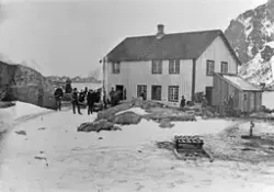 Bildet viser en del menn som står utenfor et hus. En slede i