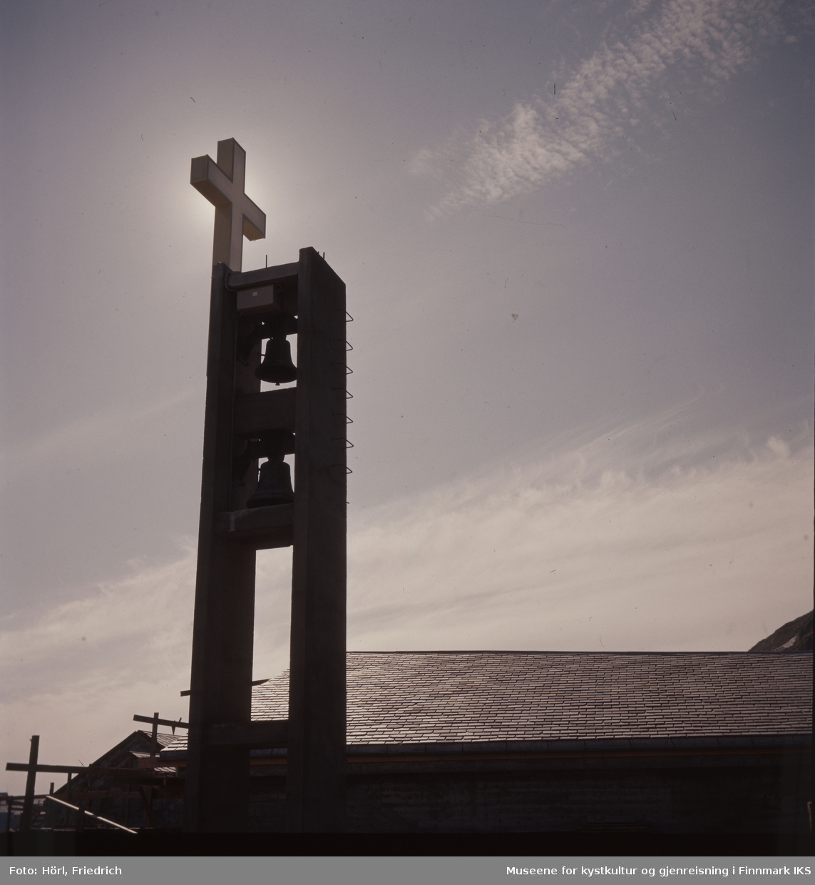Kirketårnet til Den katolske kike St. Mikael i Hammerfest med kors og klokkene er fotografert i motlyset. Tydelig ser man kirketaket som er tekket med rektangulært takskifer. Til venstre for bygget står det fortsatt noe stilas.