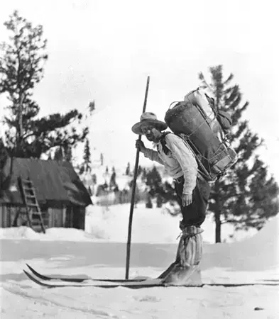 På ski iført skjorte, hatt og store gamasjer, en stav og en stor sekk og bør på ryggen