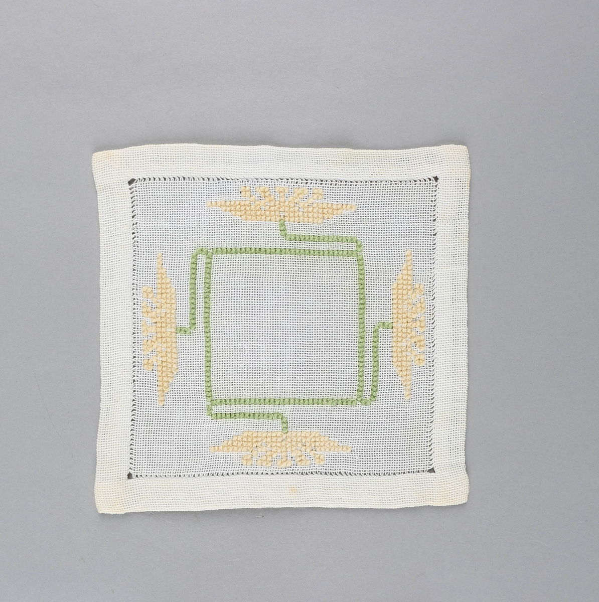 Tilnærmet kvadratisk dekkebrikke i hvitt tekstil med brodert, grønt kvadrat med fire stilker i sidene med gule blomster i endene.
