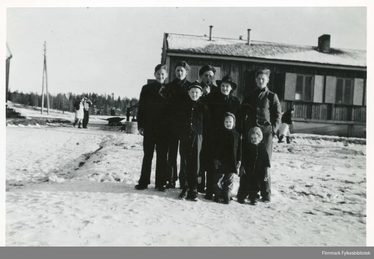 Familien Alexandersen ble evakuert fra Hammerfest i 1944 til Trøgstad i Østfold. Her er de oppstilt utenfor huset i Trøgstad fordi bøndene skulle komme og sjekke hvor mange de var, hvor sterke de var med tanke på å kunne settes i arbeid. Åtte familiemedlemmer på utstilling så alle kunne bedømme hvor mange de hadde plass til og hvor mye hjelp de kunne få av familien. Bildet er tatt i november 1944. 