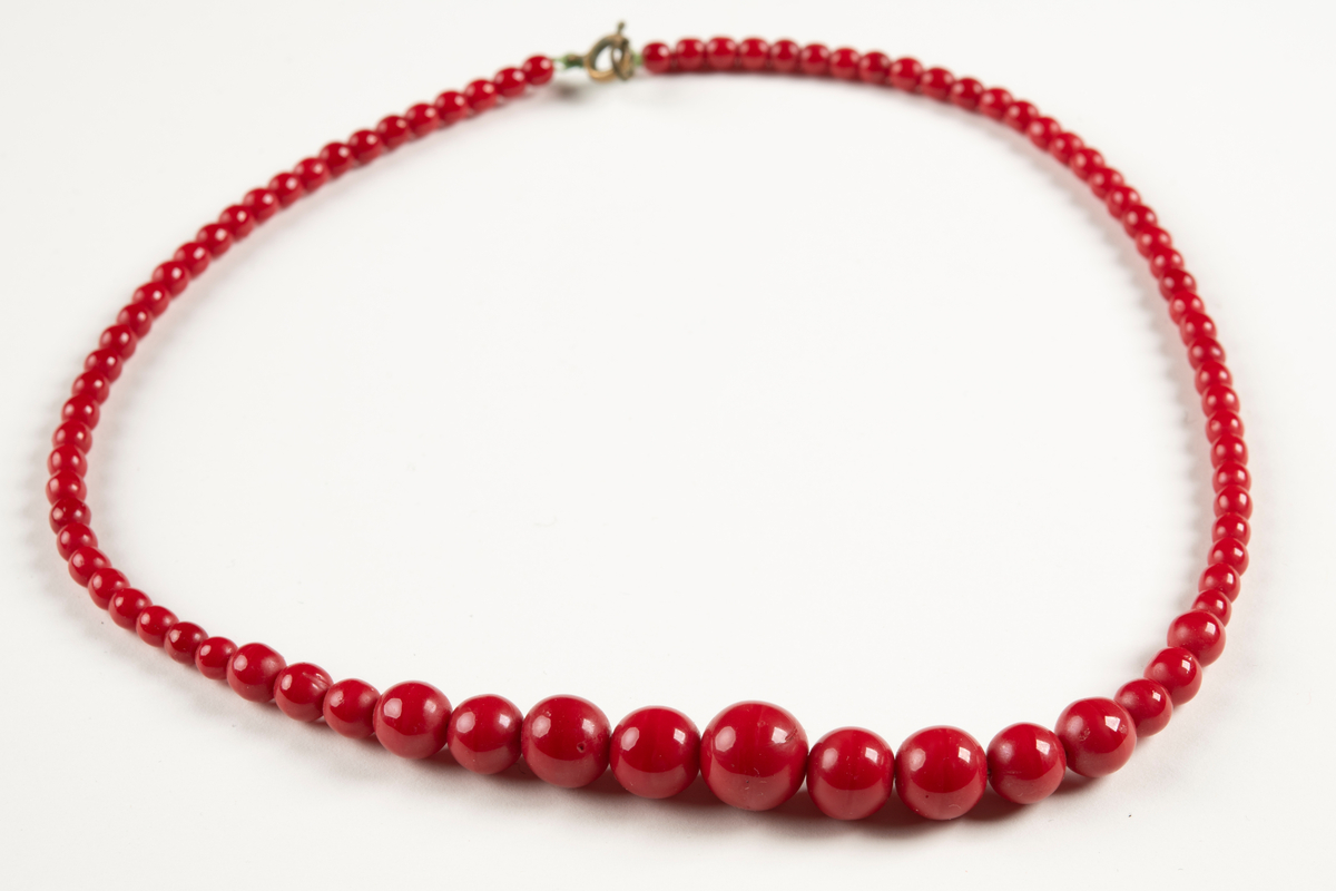 Kort halsband av röda plastpärlor trädda på en nylonlina. En stor pärla i mitten och sedan successivt mindre pärlor på ömse sidor fram till springring och ögla i metall som utgör halsbandets lås. Nylonlinan knuten i låset.