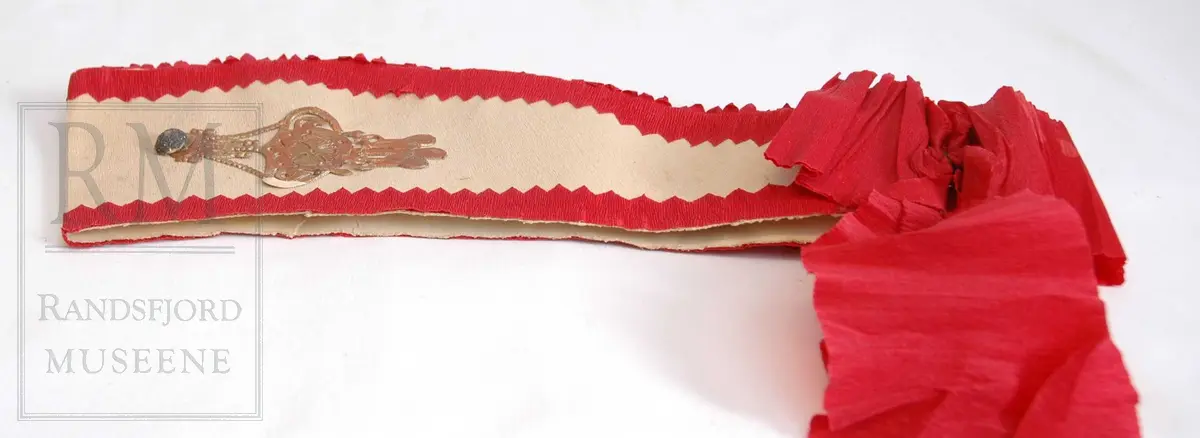 Hodeplagg for sjah e.l. Krepp på kartong. Silkepapir. Rød kreppsløyfe bak. Gyllen mansjett på sidene