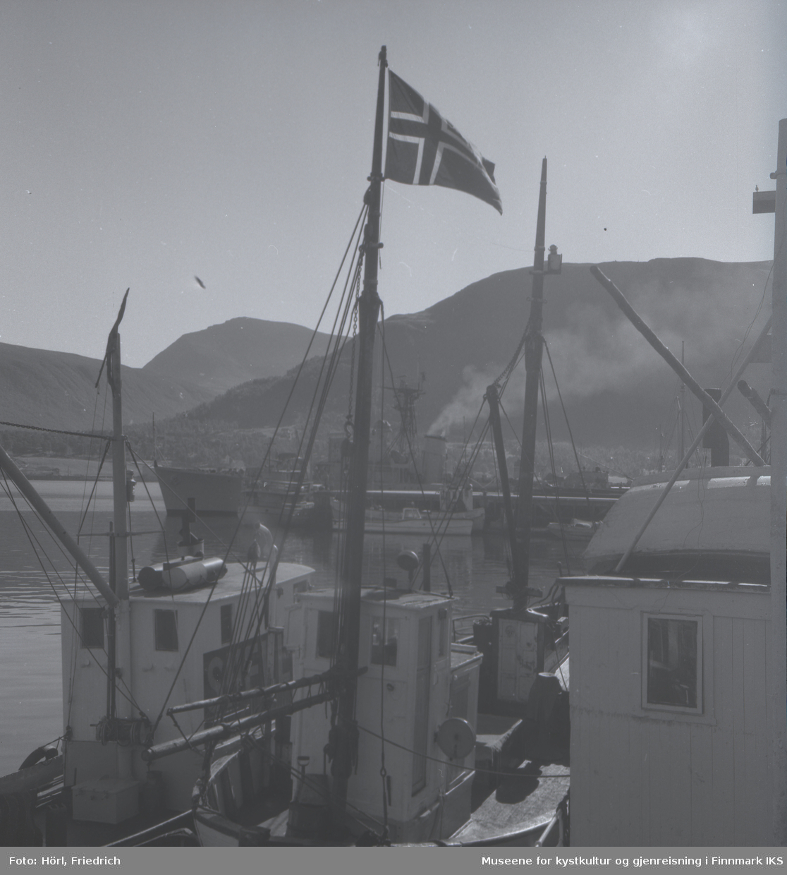 Båter av ulike typer ligger til kais i Tromsø. I forgrunnen ser man en fiskebåt med det norske flagget heist i masta. I bakgrunnen ser man Fløyafjellet.
