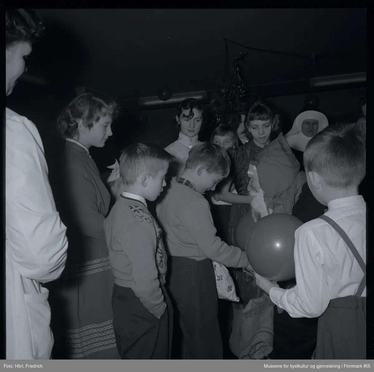 Den katolske menigheten i Hammerfest feirer juletrefest i 1957. En gutt har fått en gave fra julenissen som snakker til ham mens han slår ned blikket og ler. Andre barn og voksne står rundt dem og følger med.  
I bakgrunnen ser man juletreet og luftballonger.