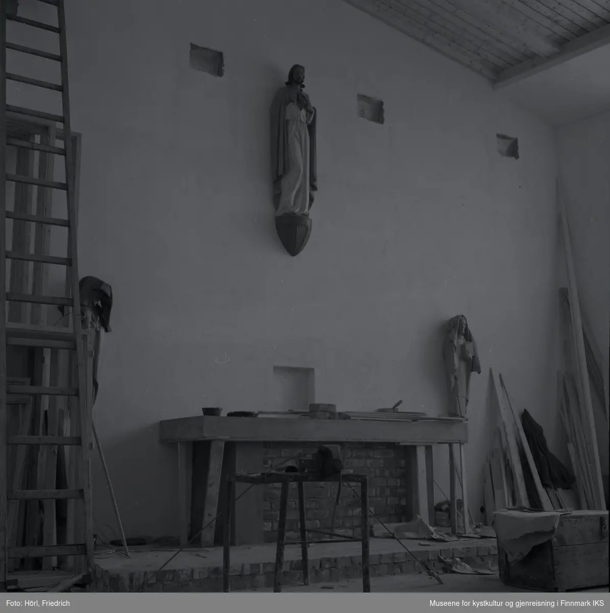 Interiøret av den katolske kirka St. Mikael i Hammerfest bygges i 1958. I motsetning til senere, er det montert en skulptur av Jesus Kristus ovenfor alteret. Senere ble det plassert et krusifiks der. Mosaikken på fronten av alteret, som ble utført av Karl Manninger, er ikke kommet på plass enda. Til venstre for alteret er det montert enn skulptur av Maria med barnet, til høyre vises Josef. Skulpturene, som er skjært av tre, er avdekket med tepper. Til venstre i bilde står en lang stige og til høyre noe mer byggemateriale lent mot veggen.