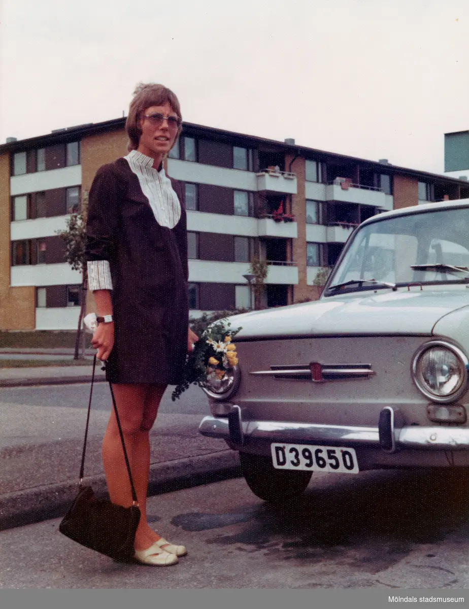 Karin Pettersson (gift Hansson), klädd i brudklänning och med en blombukett i handen, står brevid sin Fiat 650 utanför bostaden på Havrekornsgatan i Bifrost, 1973-09-29.