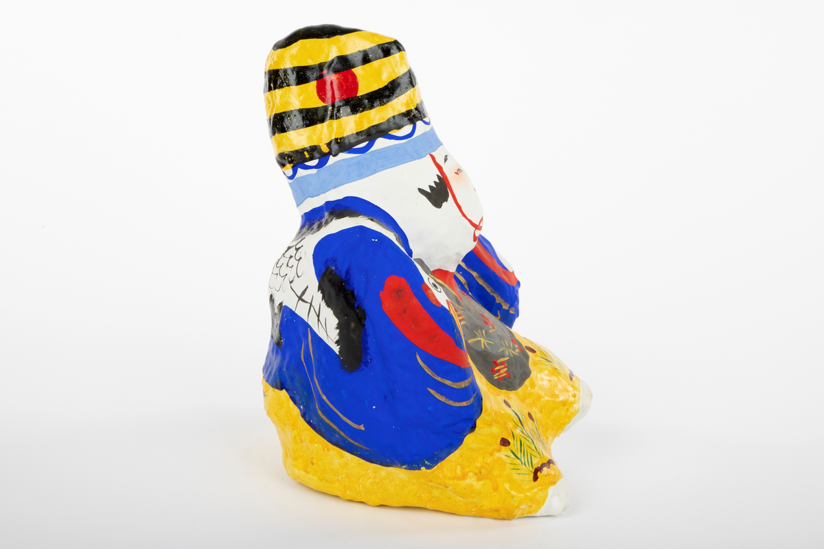 Mannsdukke i pappmasjé med stort hode og liten, kompakt kropp. Figuren har gul bukse, grå vest, blå jakke og en gul- og svartstripete hatt. På dukken er det malt flere lykkesymboler, blant annet en trane på ryggen.