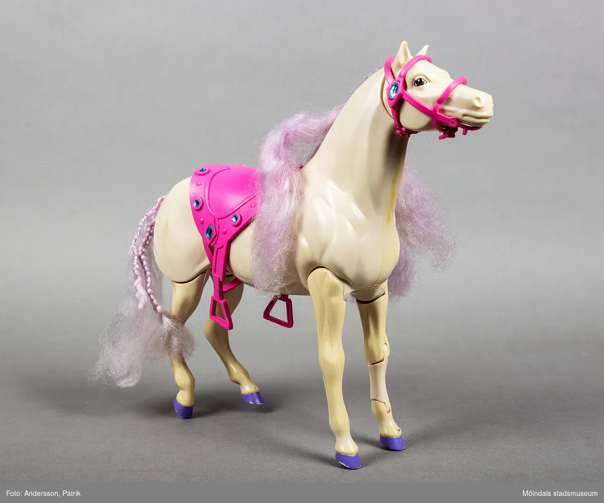 Vit leksakshäst i plast med lila man och hovar. Benen är ledade och rör sig när man leker med den. Hästen har sadel och betsel i rosa plast med grönblåa ”ädelstenar”.