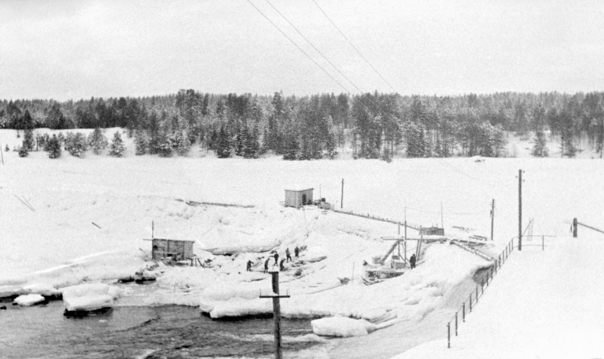 Skjefstadfossen i Heradsbygda i Elverum i Hedmark, fotografert fra den østre elvebredden.  Dette fotografiet ble tatt i mars 1937, på et tidspunkt da mesteparten av elveløpet var islagt og snødekt, og da vannføringa i elva var meget beskjeden. Den beskjedne vannføringa var årsaken til at man, til tross for is, snø og et ubekvemt arbeidsklima, utførte reparasjoner på den 312 meter lange terskeldammen som hadde gått i en bue over elveløpet siden Skjefstadfossen kraftverk ble bygd i 1909-1910 på denne årstida. På bildet skimter vi den buete damkrona med et slags stillas og ei lita bu med pulttak. Også på isen i fallsona under dammen var det bygd et lite skur med pulttak. Mellom dette og dammen skimter vi et arbeidslag som hakket is, antakelig med sikte på å frilegge konstruksjoner som skulle repareres før vårflommen kom.