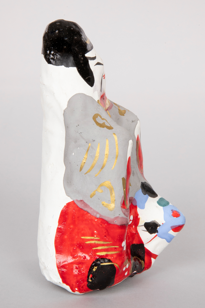 Figur i pappmasjé som forestiller en stor rytter på en liten okse. Mannen har på seg rød og grå kappe med gullbånd. Japanske tegn på baksiden av figuren.