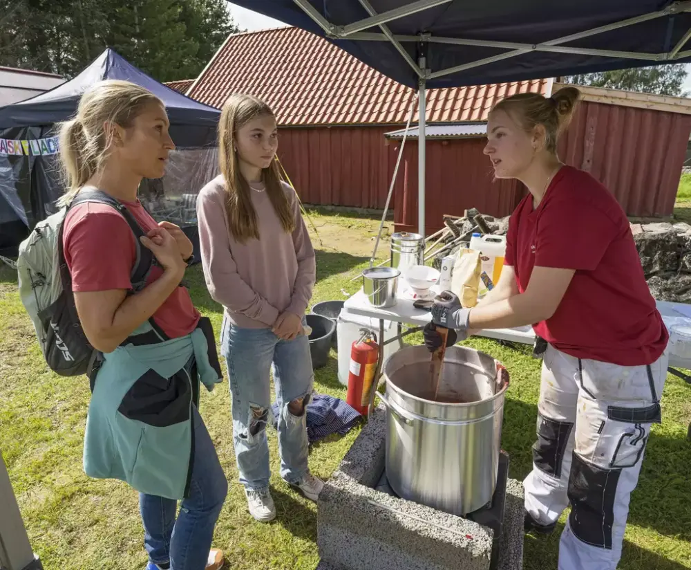 En ung kvinne som er tradisjonshåndverker blander komposisjonsmaling i en kjele, og forklarer til to kvinner som ser på.