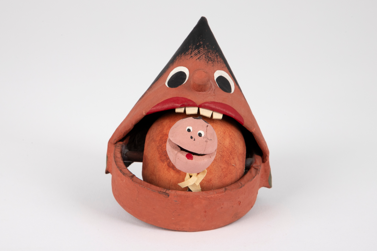 Kjegleformet, bevegelig uhyre i leire. Figuren har en brun kule i munnen, hvite tenner av papp og et lite rosa hode som stikker ut av munnen.