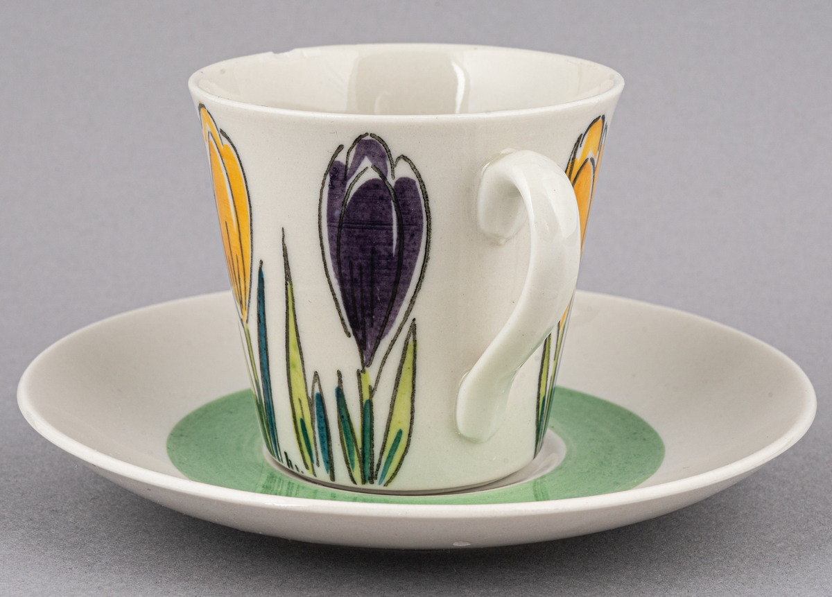 Kaffegods i flintgods med rikt handmålad dekor i form av krokusar i gult och lila, modell E. Dekoren skapad av Ellen Mortensen 1959, tillverkades till 1964. Koppen har två nagg vid kanten.