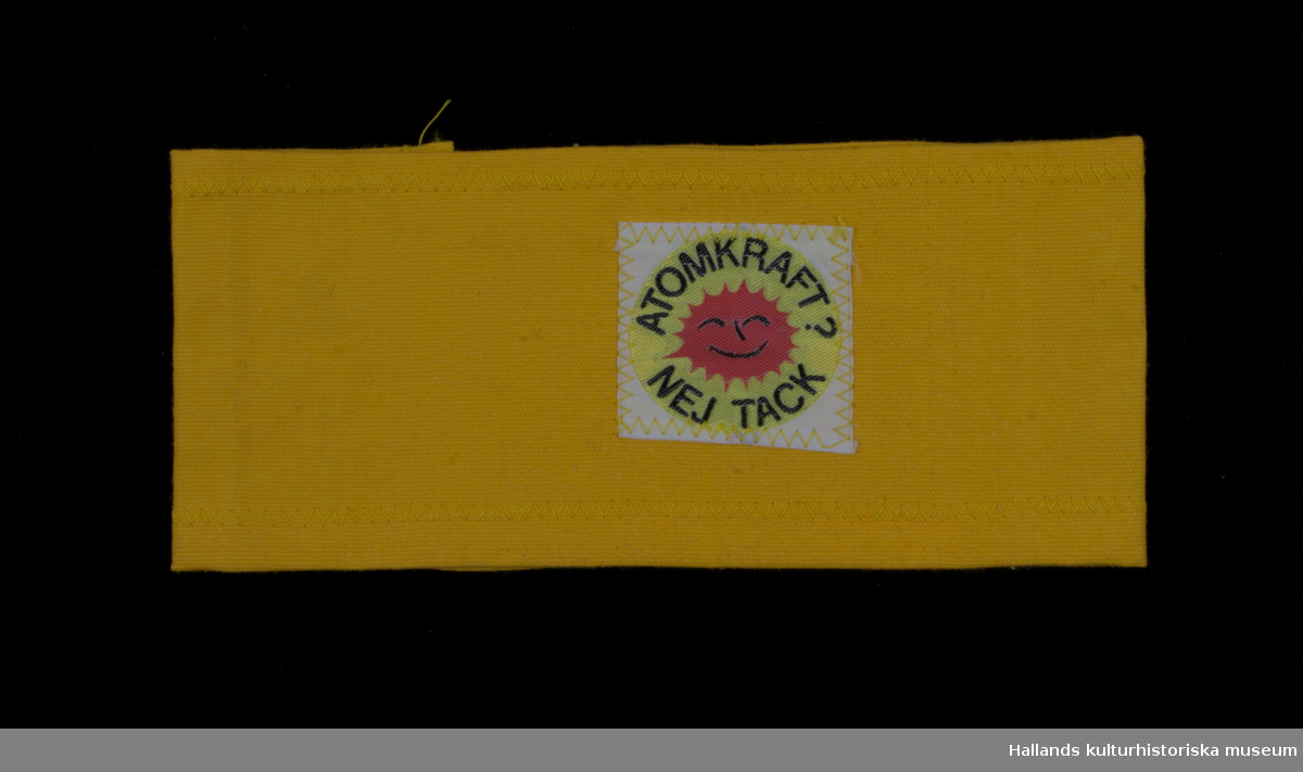 Armbindel av gult bomullstyg. Påsydd antikärnkraftssymbol: gul botten med röd skrattande sol samt svart text: "ATOMKRAFT? NEJ TACK".