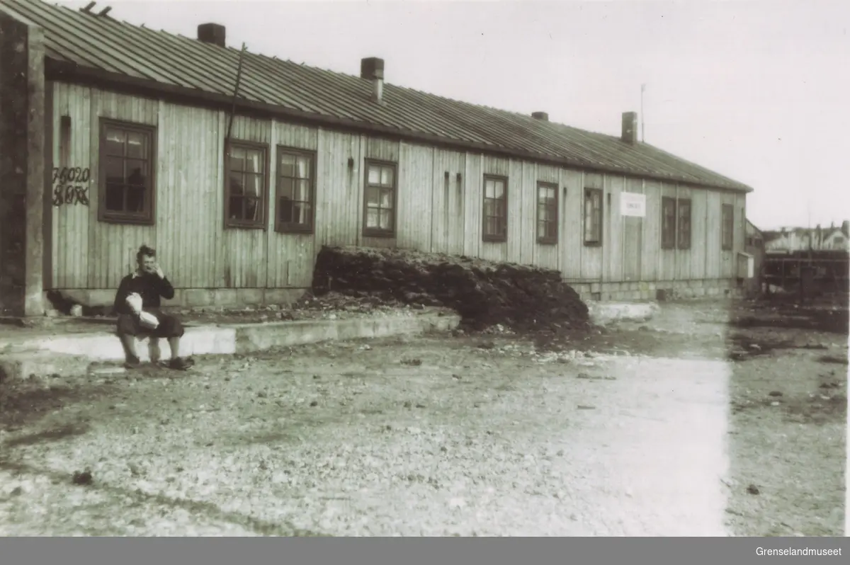 Avbildet i svart-hvitt er gjenreisningsadministrasjonens brakke i Kirkenes som lå der rådhuset ligger i dag. En person sitter på fortauet foran brakkebygget.