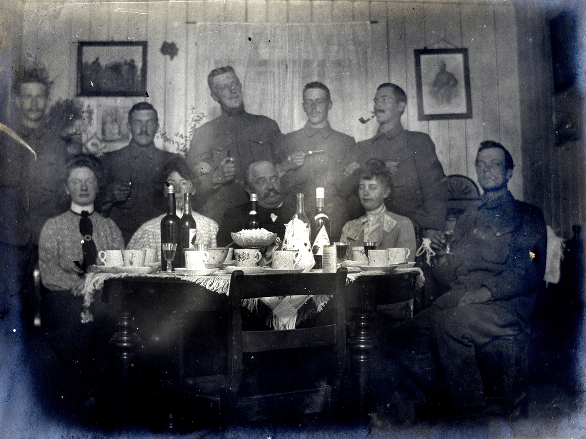 Soldater og kvinner sitter og står ved et bord med flasker og kopper på.
Fest på Askhuset 1905.
Bilde er fra fotoalbum GM.036887.