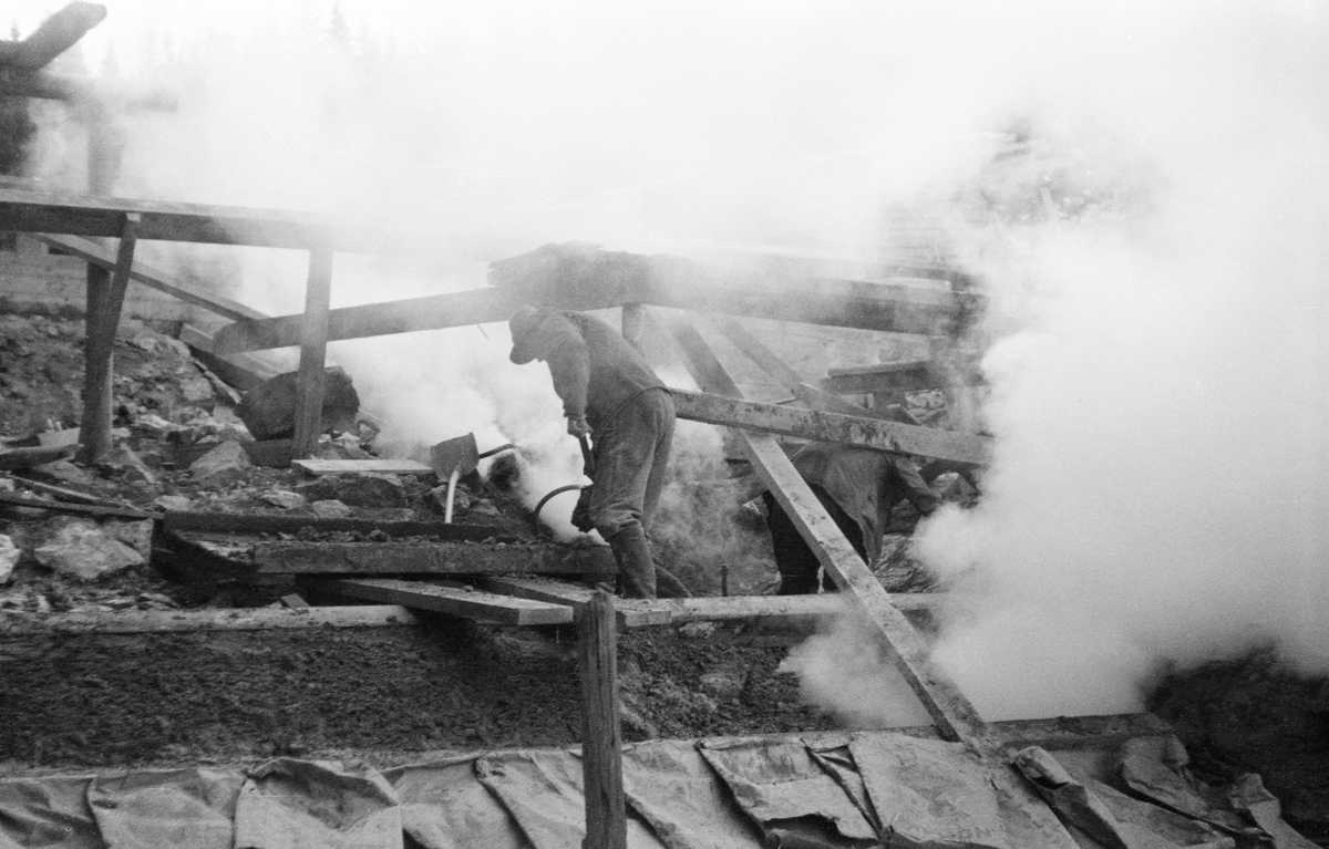Fra bygginga av en ny dam ved Sølvstufossen, øverst i Ågårdselva i Tune i Østfold, som er en del av Nedre Glommas vestre løp. Anleggsarbeidet startet vinteren 1936, i en periode da det knapt rant vann i elveløpet. Fotografiet er tatt fra elveløpet, antakelig der man skulle bygge dammens bunnløp. To menne arbeidet i ei sky av røyk eller støv fra et pressluftbor. Ågårdselva er et cirka fem og en halv kilometer langt vassdrag som renner fra den nordvestre enden av Isnesfjorden (Vestvannet) i Nedre Glommas vestre løp, sørvestover gjennem berglendt terreng mot innsjøen Visterflo. Høydeforskjellen mellom Isnesfjorden og Visterflo er på bortimot 25 meter. Ågårdselva har tre fossefall, det øverste her ved Sølvstu, deretter ved Valbrekke og nederst ved Solli. Behovet for den dambygginga vi ser på dette fotografiet var forårsaket av selskapene Borregaards og Hafslunds inngrep i Glommas østre løp, hovedløpet, som i lavvannsperioder gjorde det svært vanskelig å få tømmer som skulle til bedrifter nedenfor Sarpsfossen via Mingevannet, Isnesfjorden og tømmertunnelen til Eidet gjennom det trange sundet ved Trøsken. Dette forsøkte man å løse ved å heve vannspeilet i Isnesfjorden med en dam som skulle plasseres 10-15 meter ovenfor den eksisterende dammen ved Sølvstufossen. Se mer informasjon under fanen «Opplysninger».