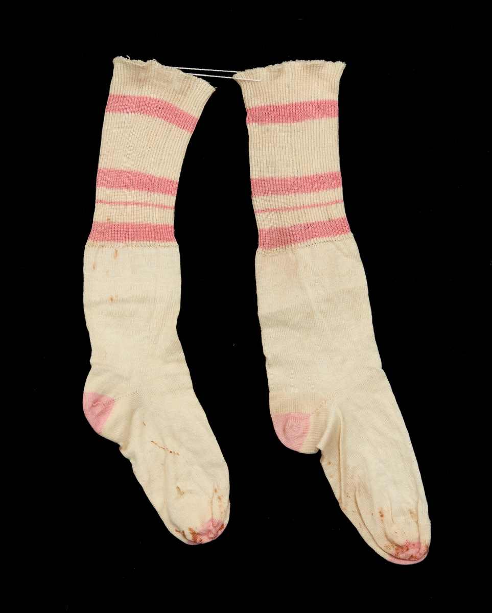 Ett par fickstrumpor, maskinstickade av vitt bomullsgarn. 12 mm av skaftet stickat med en rät och en avig maska och med tre bredare och en smal tvärgående rand i rosa. Resten av skaftet samt foten är slätstickad emd häl och tå av rosa garn. Använd 1910-tal.