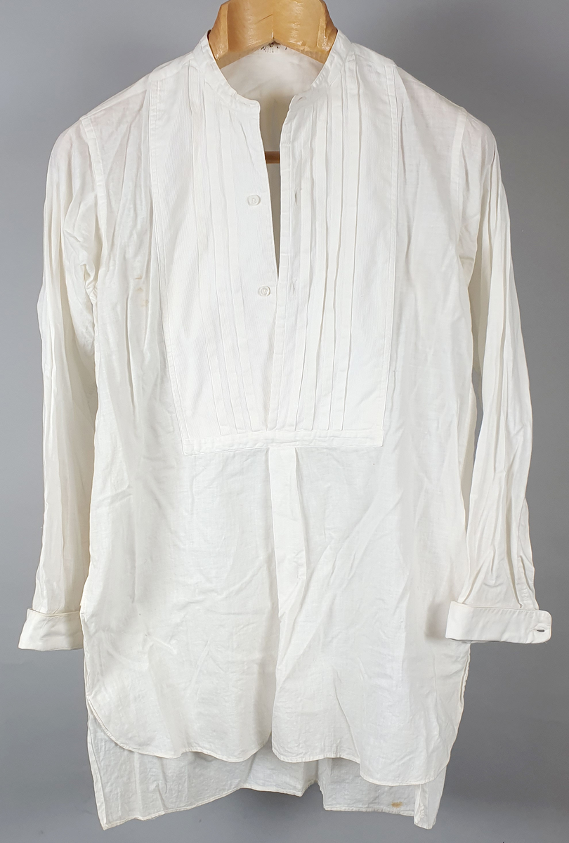 Hvit skjorte bryststykke med vertikale folder og knapper. Splitt i sidene. Til skjorten følger en løskrage.