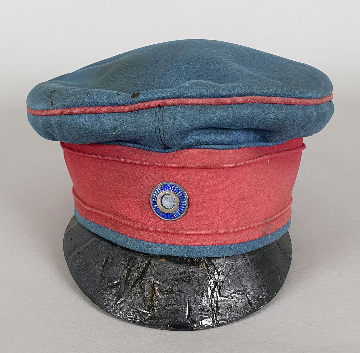 Lyseblå uniformslue med rødt bånd rundt pullen. Blå rosett midt foran.