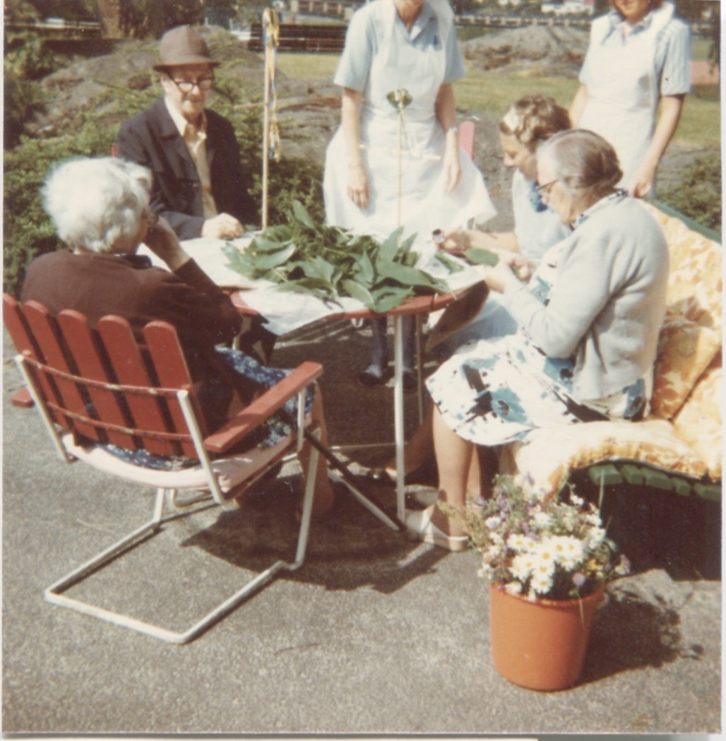 Fyra åldringar sitter runt ett bord i Brattåshemmets trädgård, 1970 - 80-tal, På bordet ligger en hög med gröna blad, som två utav kvinnorna gör något med. I bakgrunden står två sköterskor. Namnuppgifter saknas. Relaterat motiv: A4275.