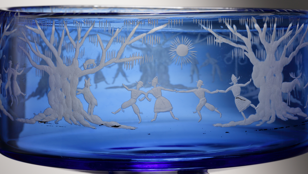 Formgivare: Gunnar Cyrén. Cylinderformad, opalblå skål med rund ofärgad klackfot. Runt livet graverat naturmotiv
med ekar, en midsommarstång och dansande och spelande människor.