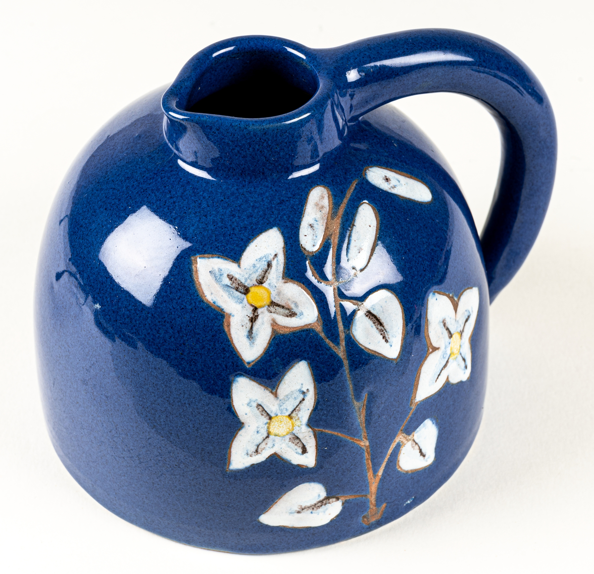 Kanna eller blomkrus i lergods, formgivet av skulptör Maggie Wibom vid hennes företag Stockholms Keramik AB, 1940-talet. Den blå glasyren fick namnet Wibomblå, kannan dekorerad med en kvist vita blommor där kvisten och konturen är oglaserad genom påläggning av vax innan glasyrbränning.