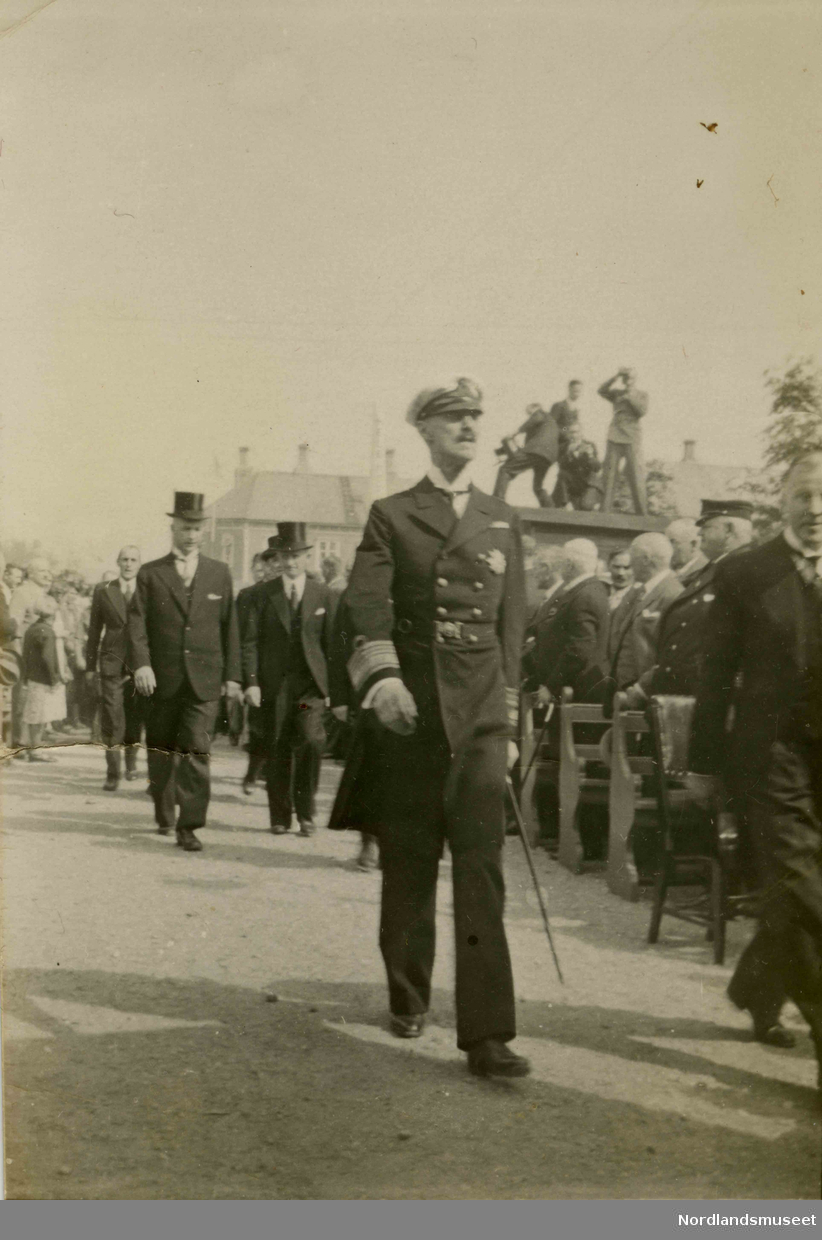 Kong Haakon VII. I bakgrunnen ser vi flere menn iført dress og flosshatt. Benker, tilskuere og fotografer sees til høyre i bildet.